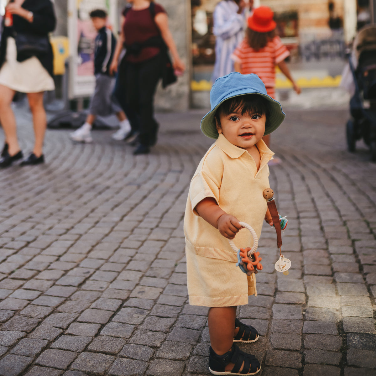 A toddler in yellow holds toys and a mushie Linen Pacifier Clip 2-Pack on a cobblestone street.