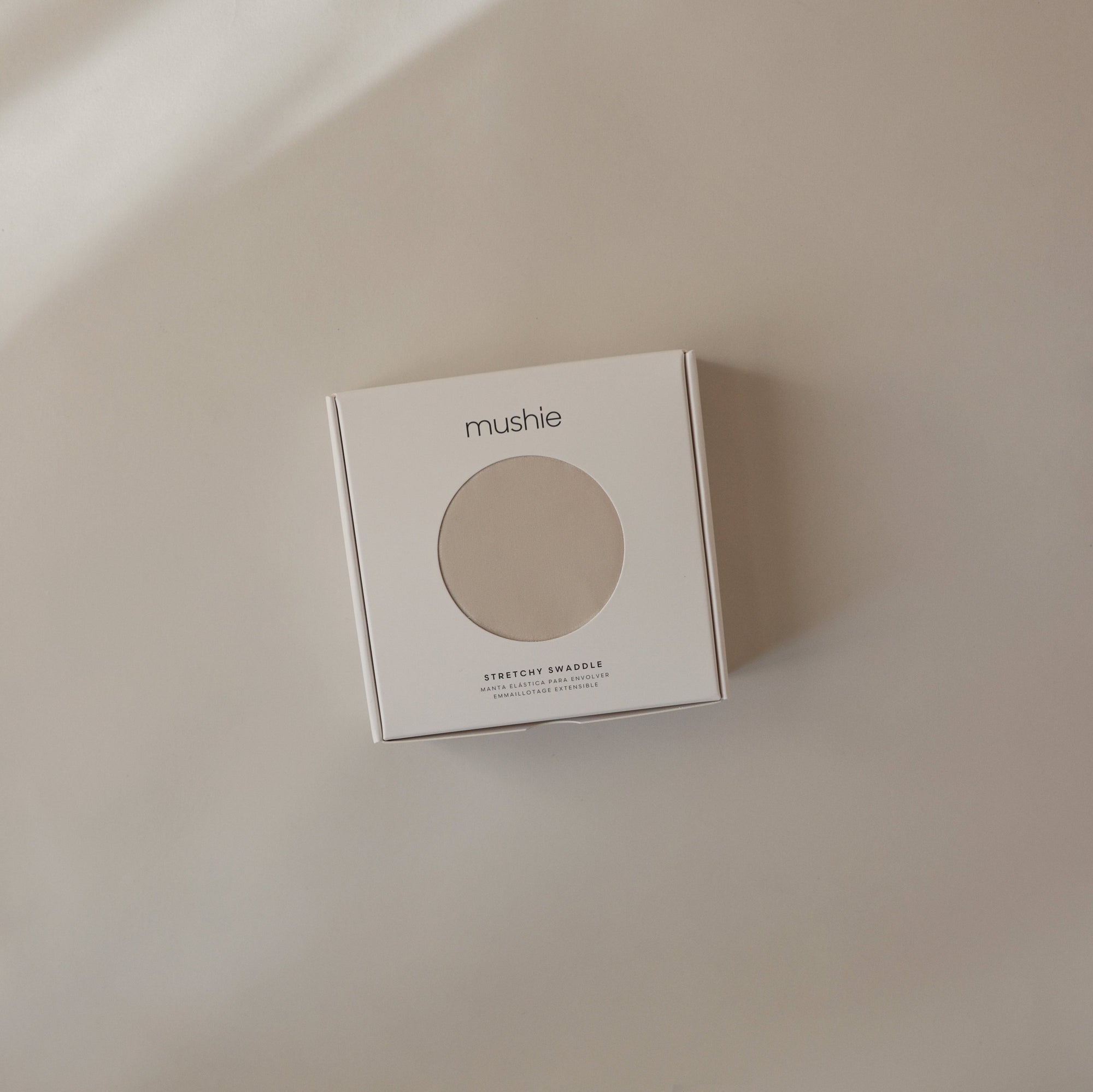 A mushie Stretchy Swaddle box with a circular window sits on a beige surface.