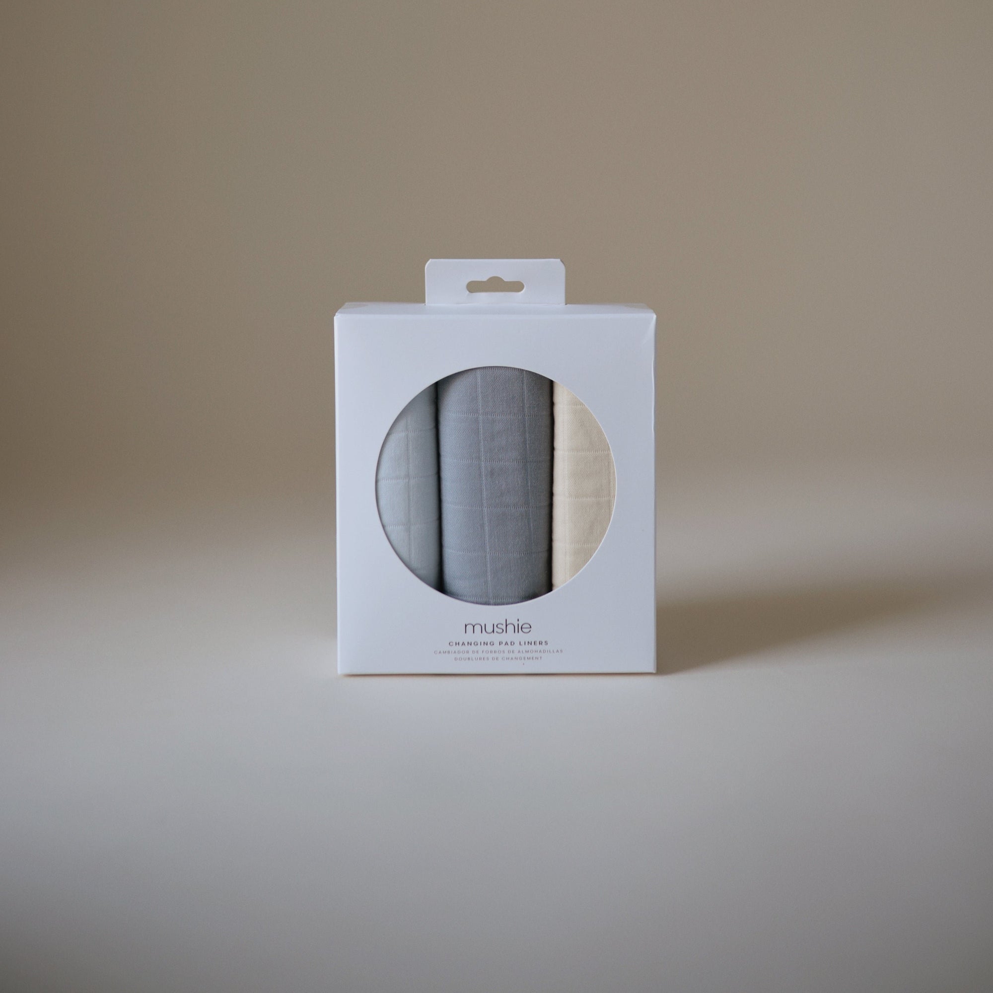 Three Mushie Changing Pad Liners, folded in a white box with a round window on plain background.
