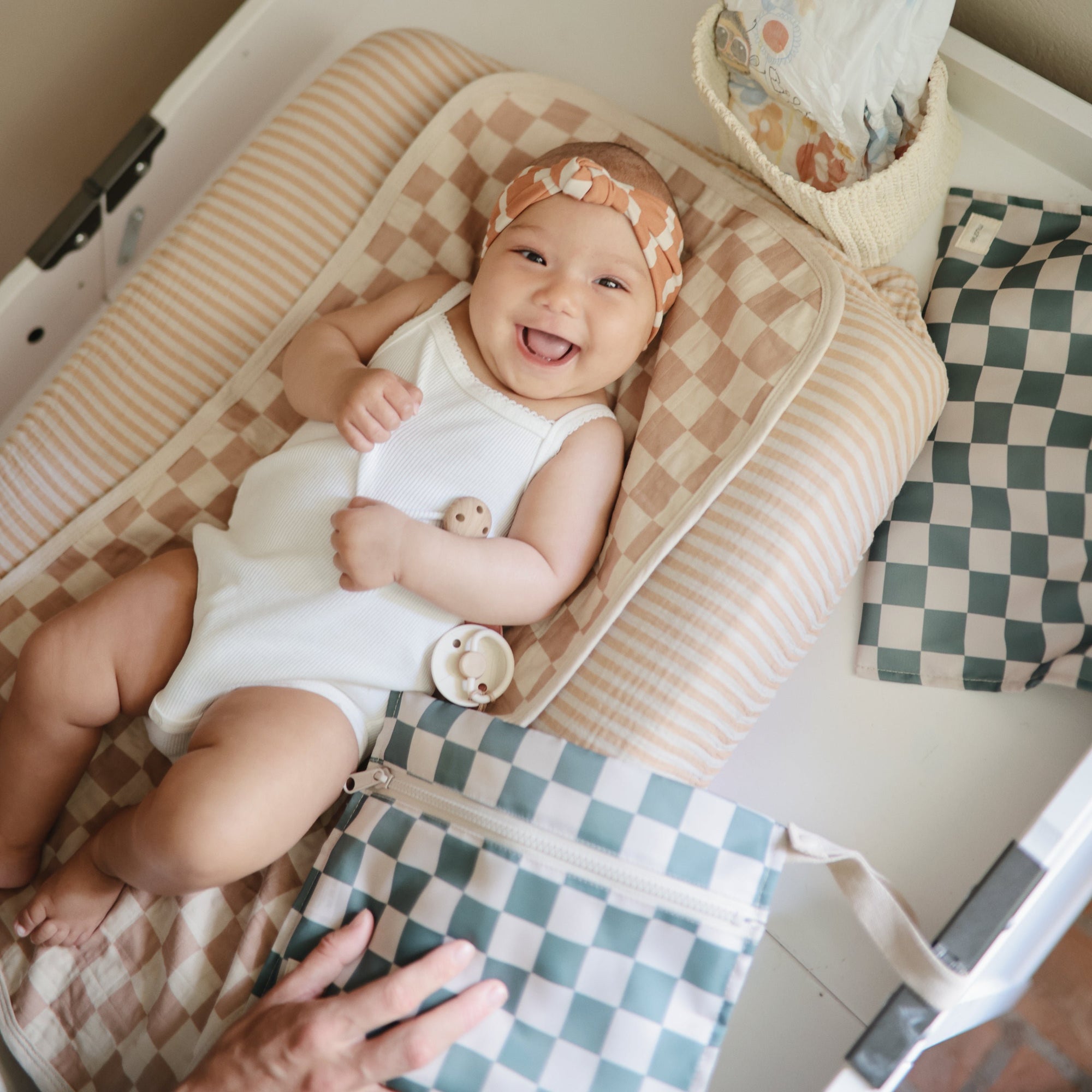 Smiling baby on Mushie Changing Pad Liners, white onesie, checkered accessories nearby.
