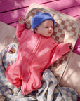 Baby in pink suit on Mushie Changing Pad Liner, blue hat, pink background.