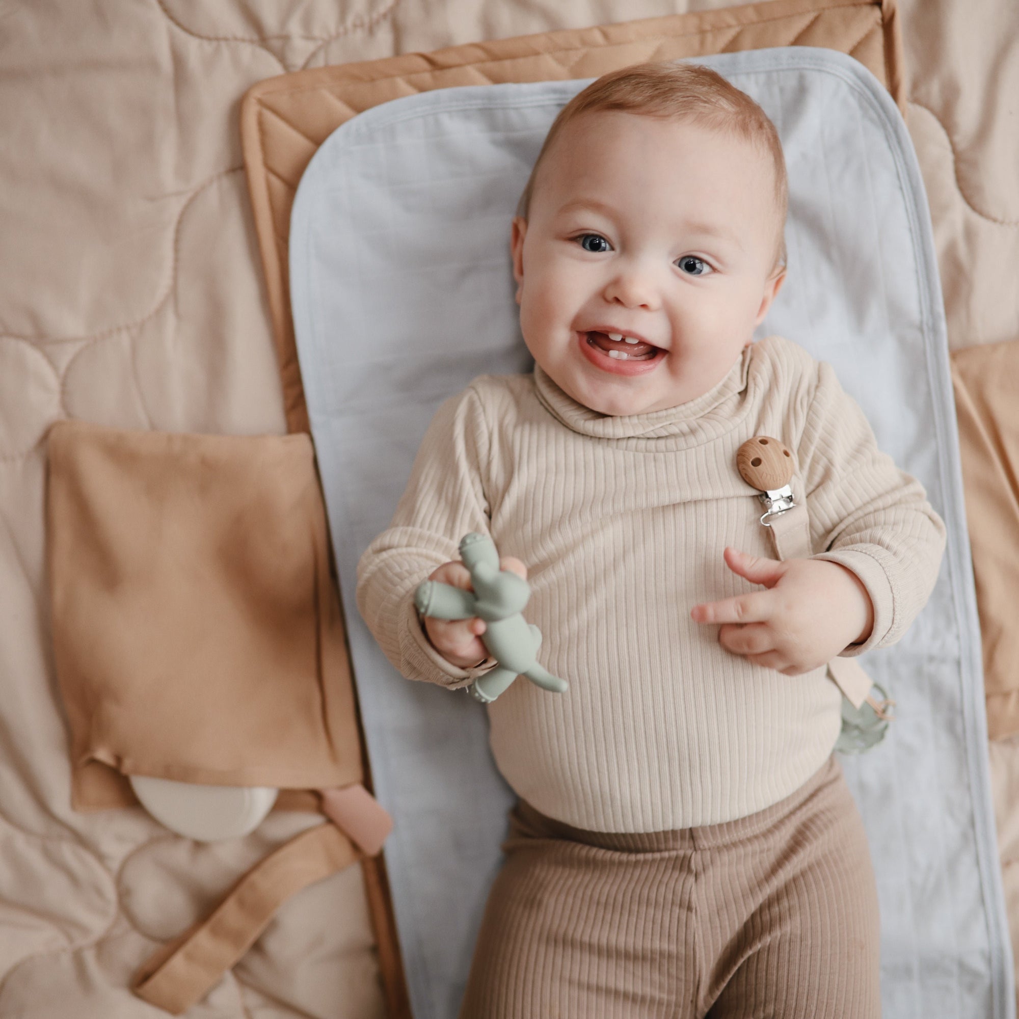 Smiling baby on Mushie Changing Pad Liner holds a toy dinosaur while lying on the mat.