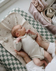 Baby in cream onesie on Mushie Changing Pad Liner, held gently near a stack of folded linens.