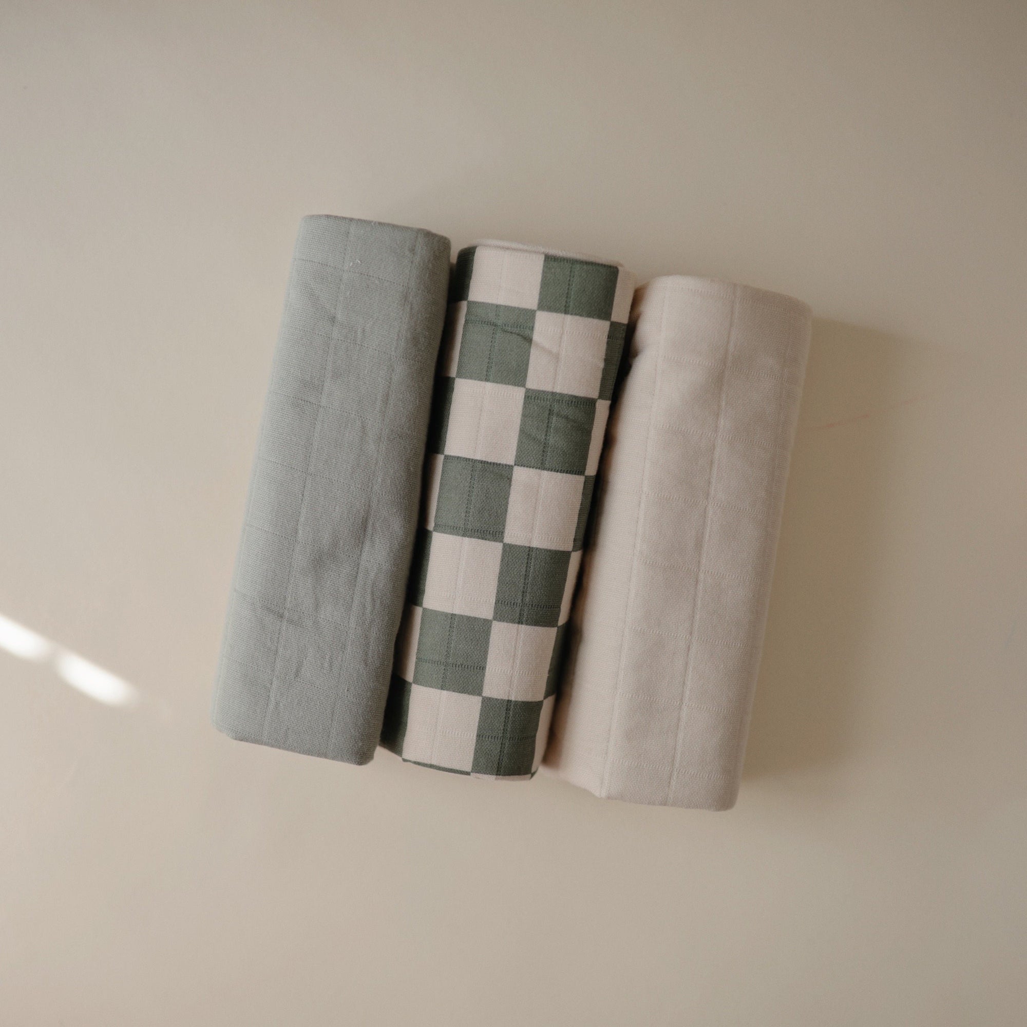 Three rolled Mushie Changing Pad Liners—green, checkered, beige—resemble organic cotton.