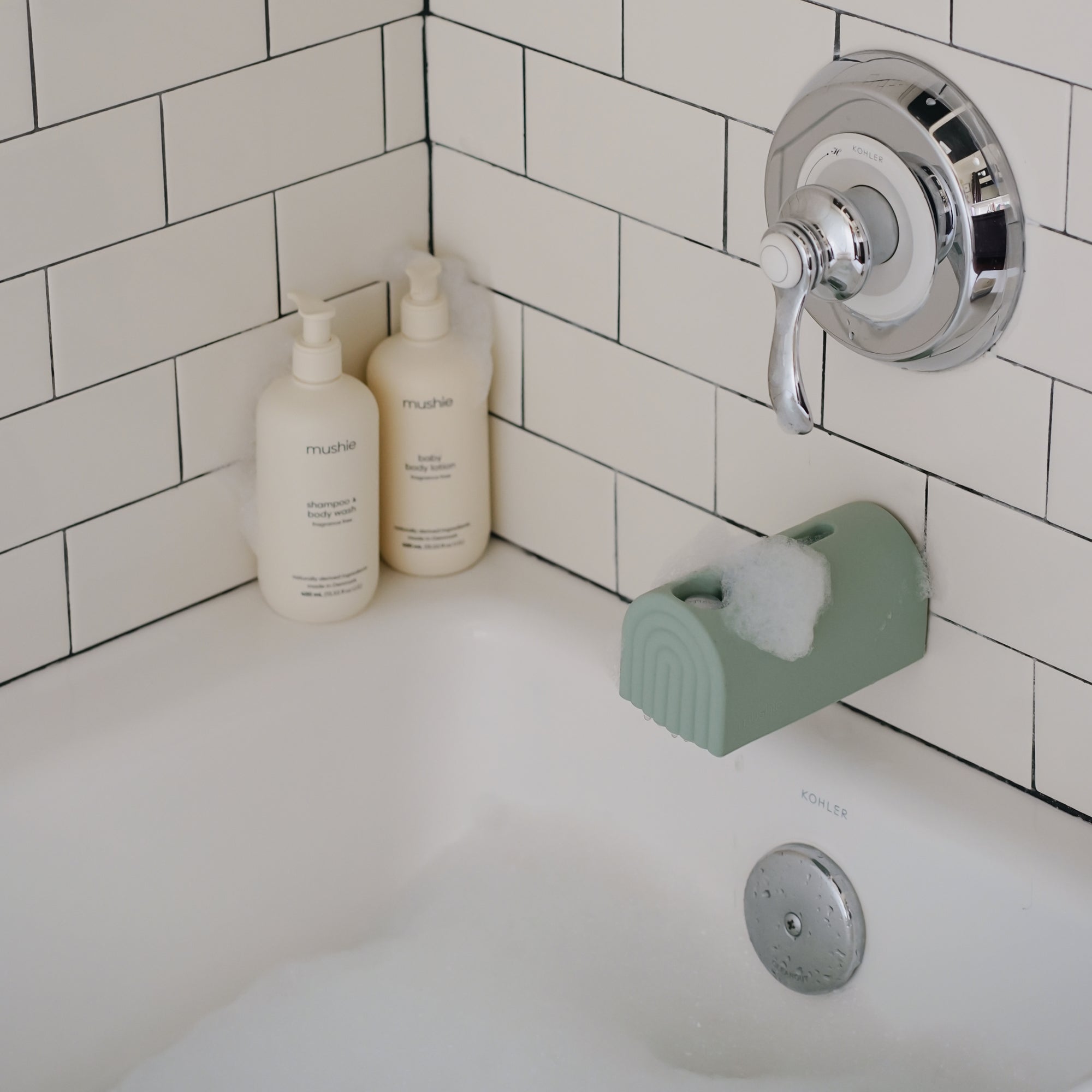 Bathtub with mushie Baby Shampoo &amp; Body Wash, white tile, and soap bottles by a green faucet cover.