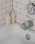 Bathtub with mushie Baby Shampoo & Body Wash, white tile, and soap bottles by a green faucet cover.