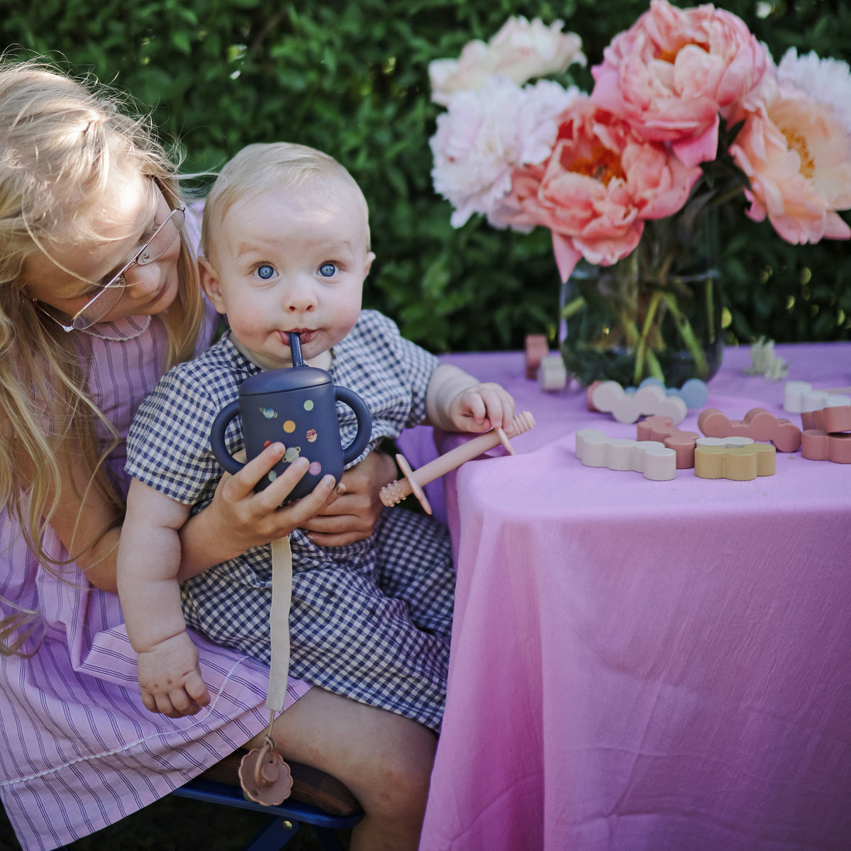 A baby with a mushie Linen Pacifier Clip 2-Pack uses a sippy cup on a girl’s lap outside.