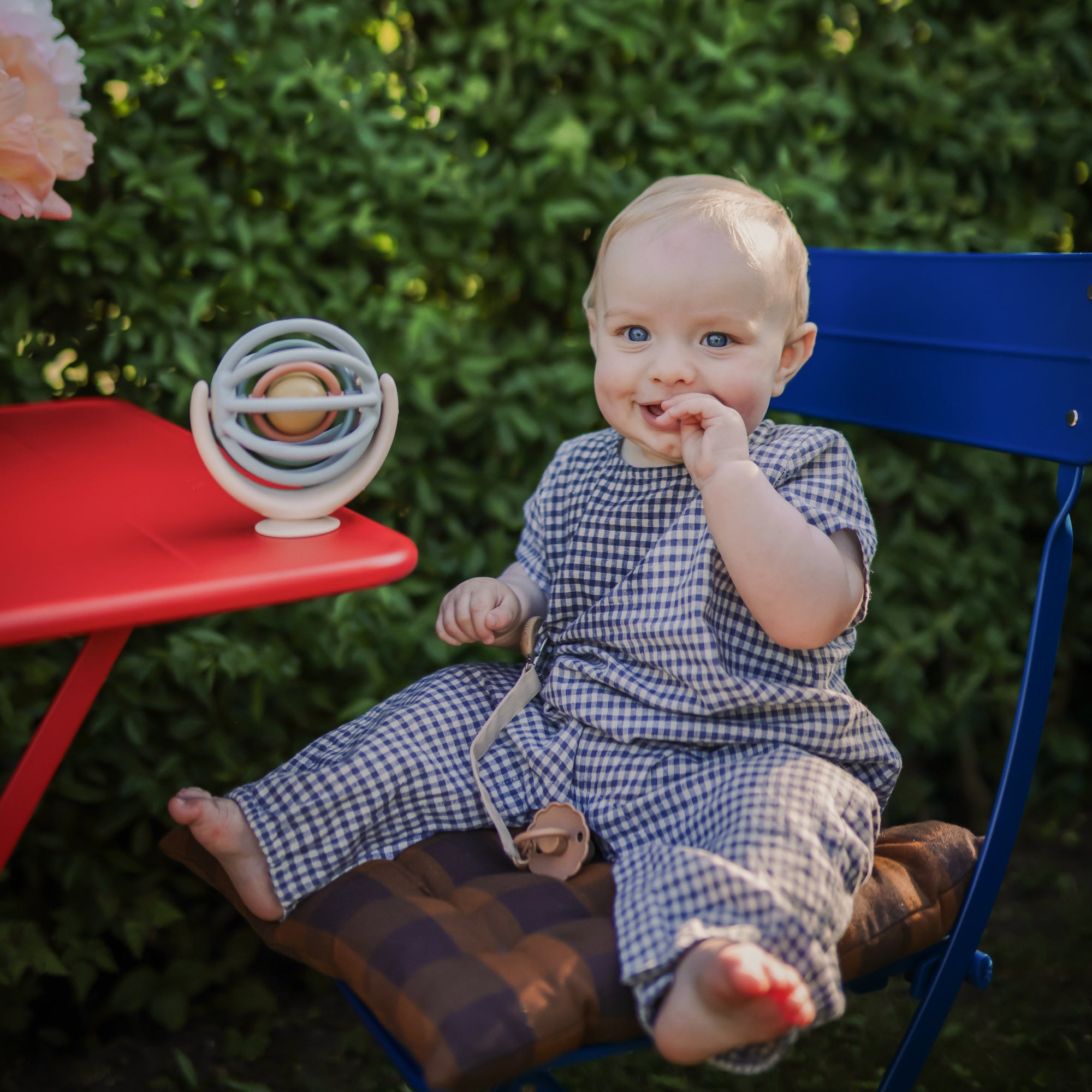Smiling baby in checkered outfit with FRIGG Daisy Silicone Pacifier by a red table outdoors.