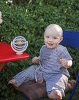 Baby with mushie Linen Pacifier Clip 2-Pack sits by red table with toy, outdoors.