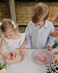 Two kids enjoy cakes and play with mushie Suction Spinner Toys at the table.