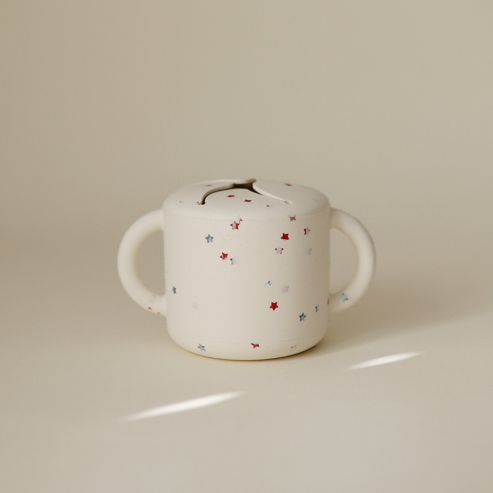 Beige mushie Snack Cup for toddlers with two handles, red and blue stars.