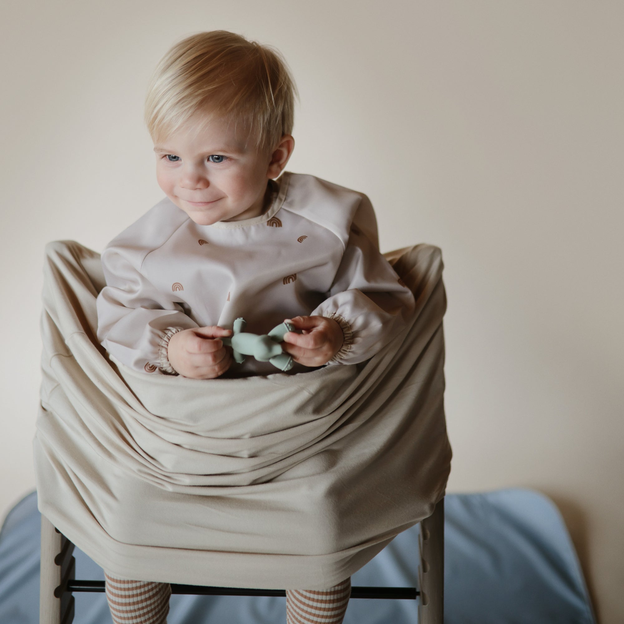 Smiling toddler in high chair holds toy, wearing mushie Multi-Use Cover and a light outfit.