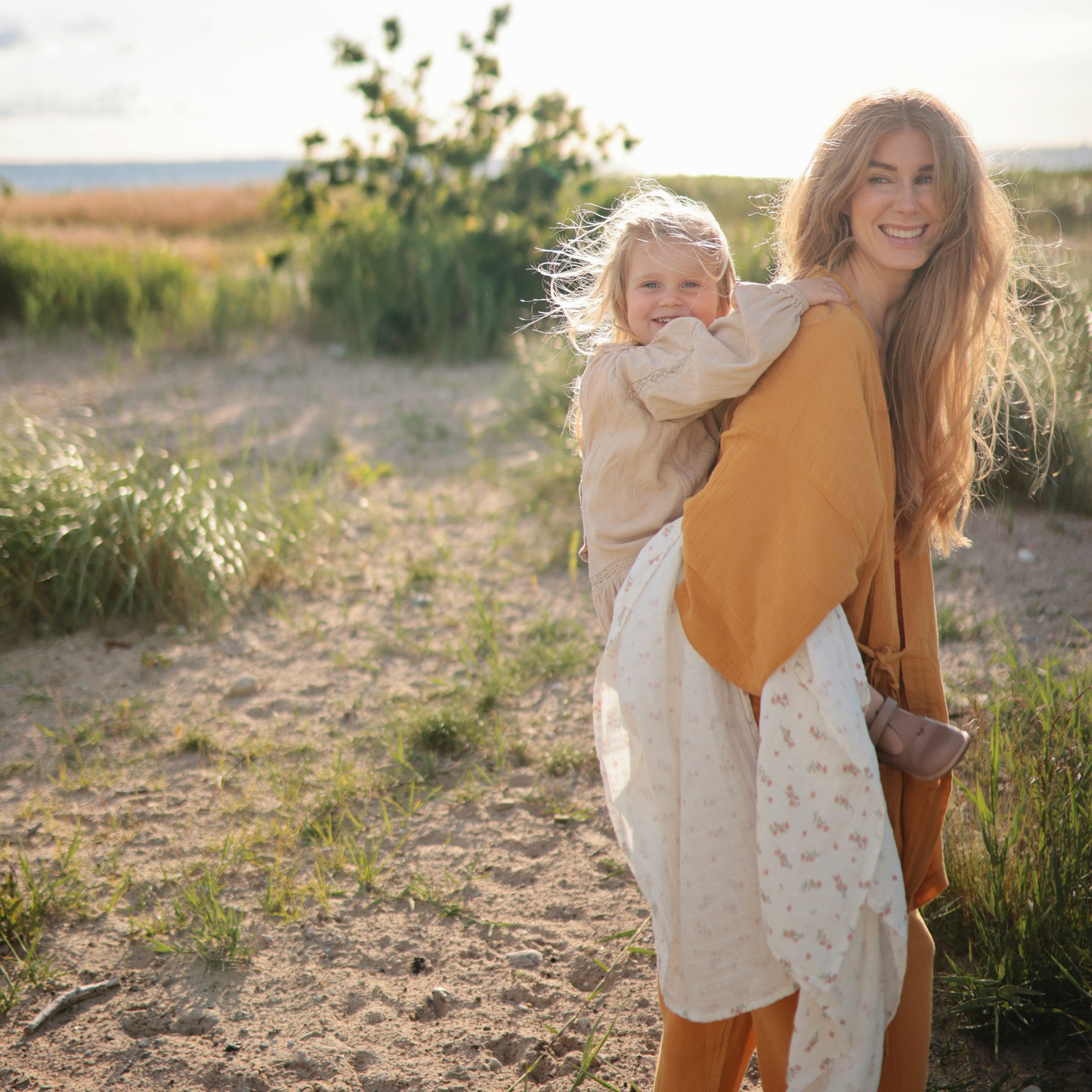 A smiling woman carries a happy child wrapped in a mushie Organic Cotton Muslin Swaddle Blanket.
