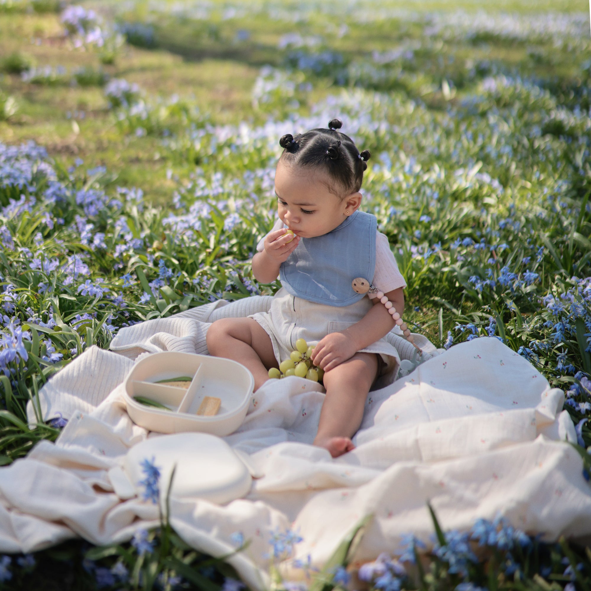 A toddler sits on a mushie Organic Cotton Muslin Swaddle Blanket, snacking outdoors in nature.