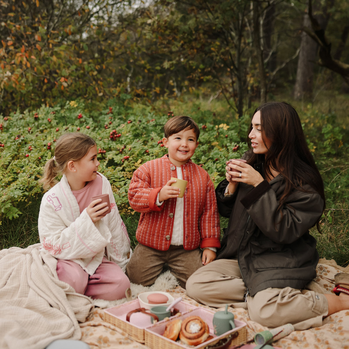 A woman and two children sit on a blanket outdoors, each holding a mushie Dinnerware Cup.