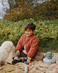 A child sits on a blanket with a small white dog and a Mushie Silicone Training Cup + Straw.