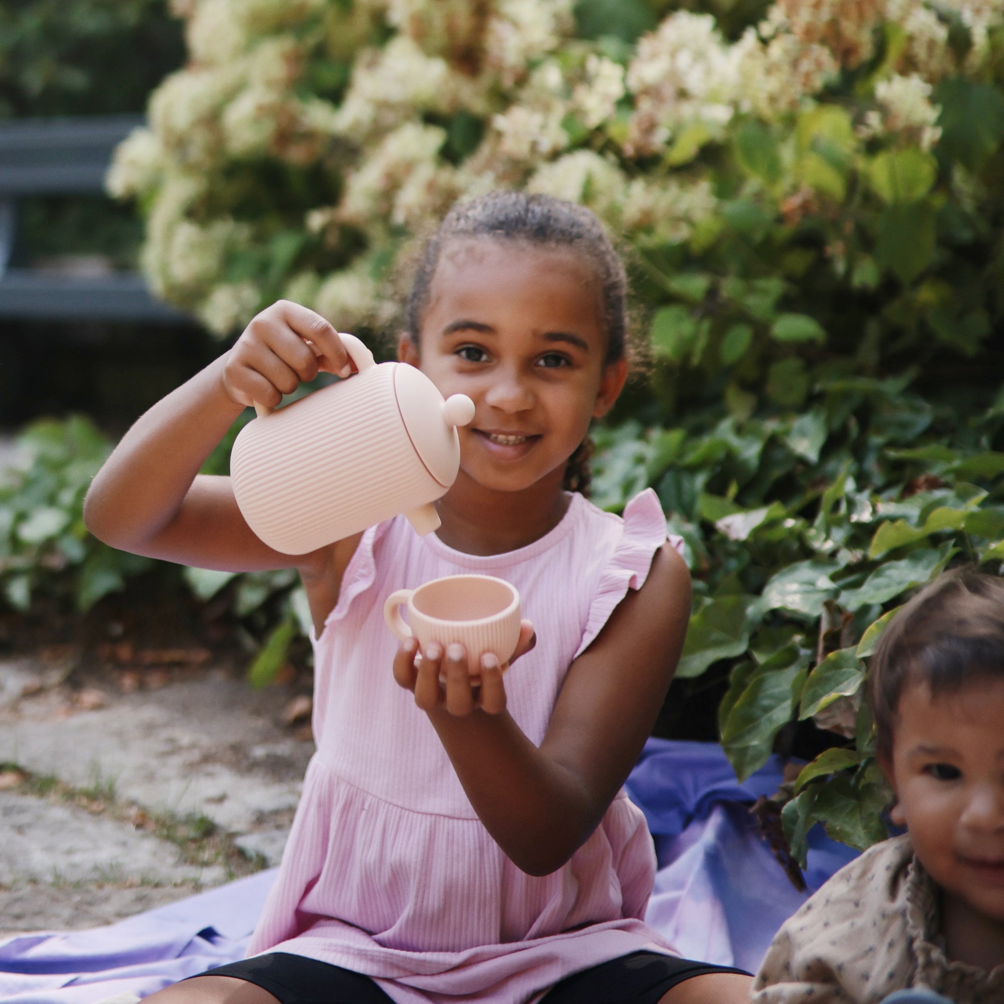 Smiling girl pours from Mushie Silicone Tea Play Set during an outdoor pretend tea party.