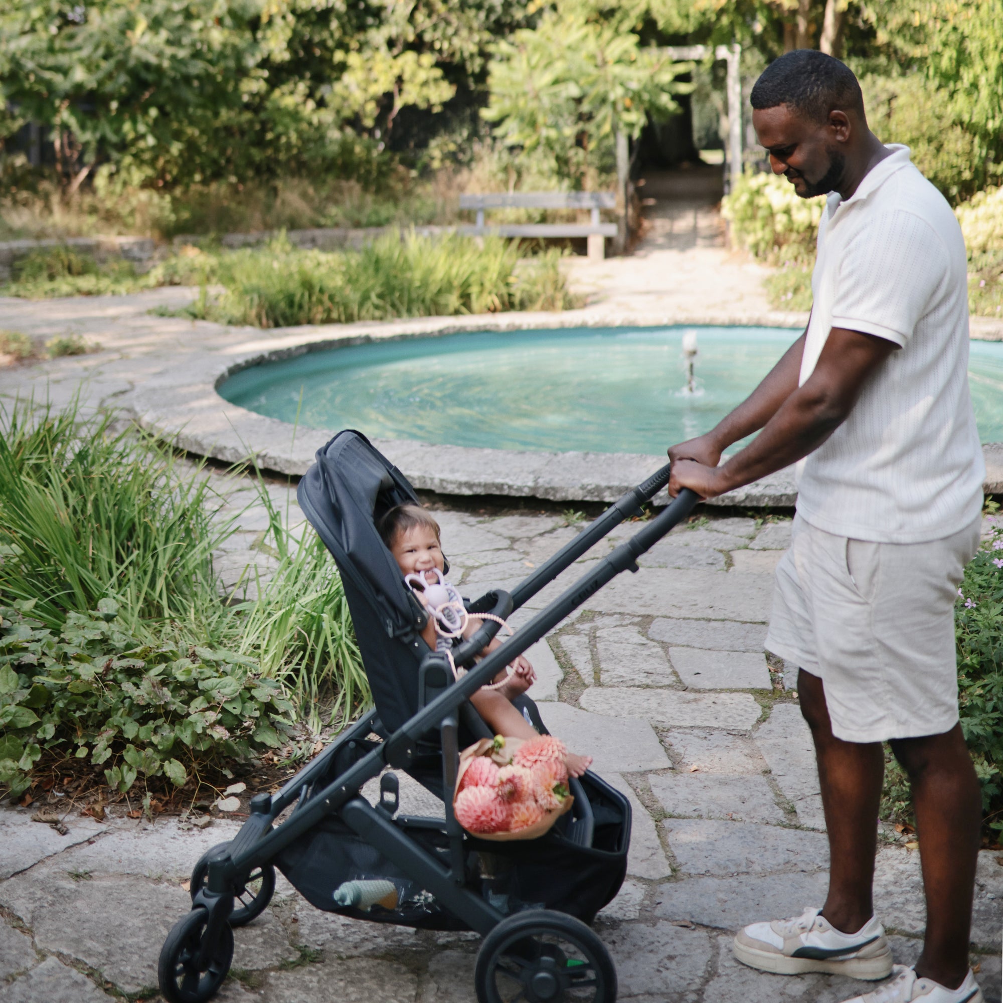 Man pushes stroller as child smiles holding mushie Daisy Rattle Teether by the garden pond.