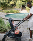 Man pushes stroller as child smiles holding mushie Daisy Rattle Teether by the garden pond.