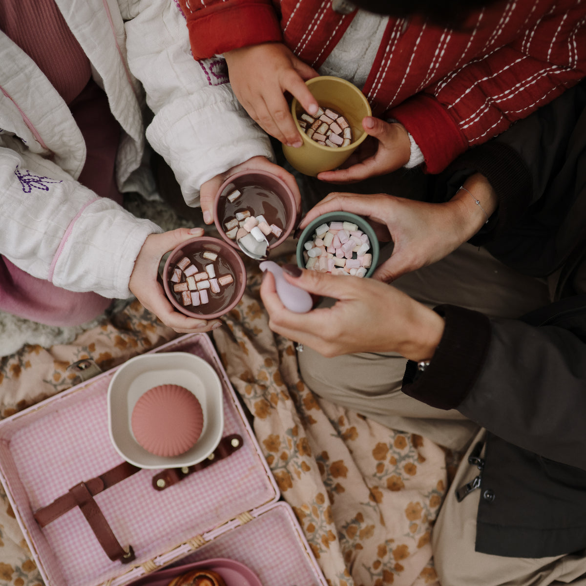 Four hands hold mushie Square Dinnerware Bowls of hot cocoa with marshmallows over a blanket.