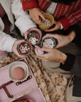 Four hands hold mushie Square Dinnerware Bowls of hot cocoa with marshmallows over a blanket.