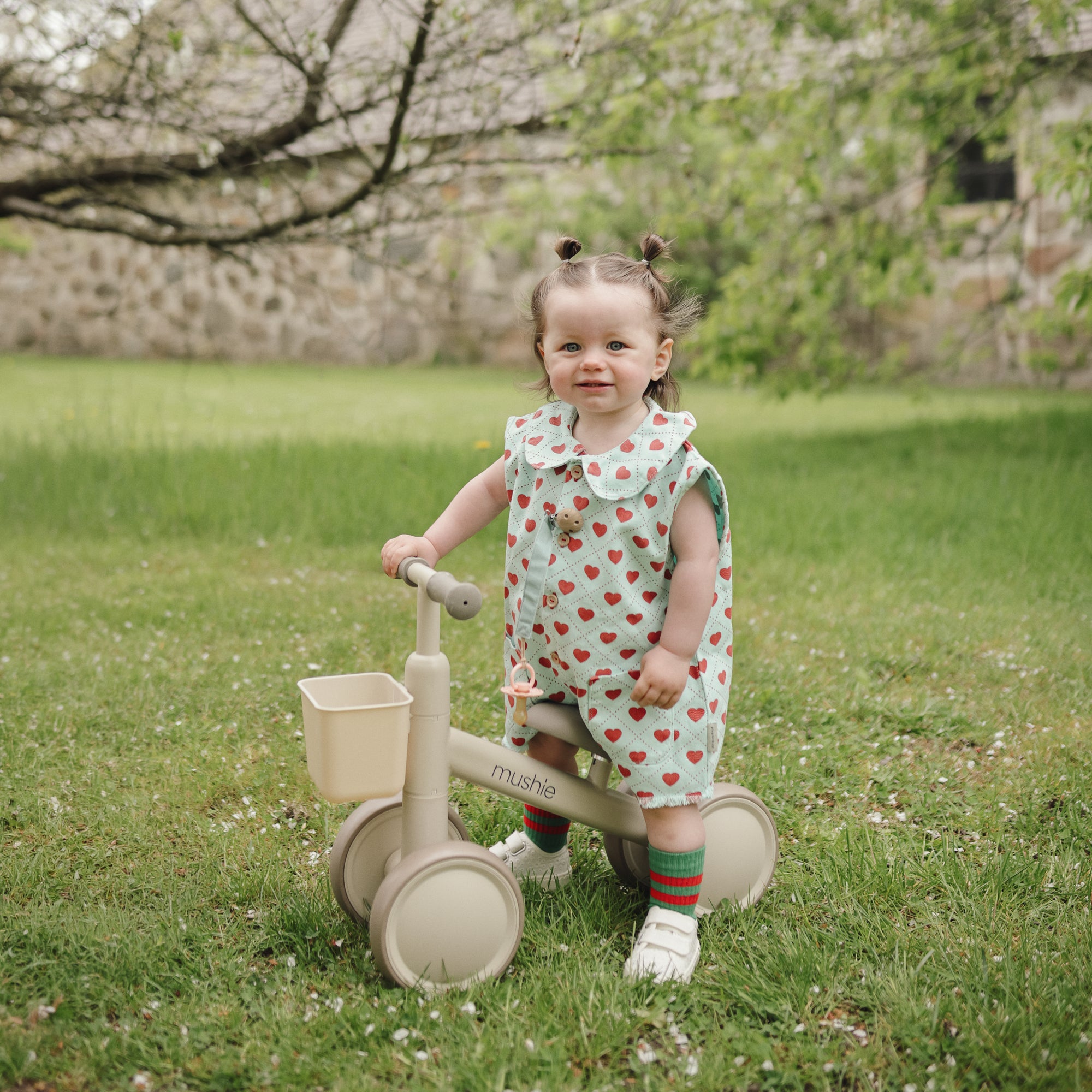 A toddler in a strawberry dress holds a mushie Linen Pacifier Clip 2-Pack by a beige tricycle.