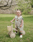 Toddler in a strawberry dress stands with a mushie Mini Toddler Bike on grass outdoors.