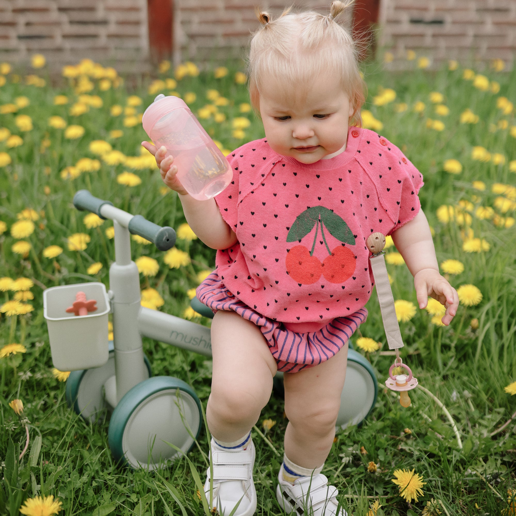 Toddler in pink cherry shirt on balance bike with FRIGG Daisy rubber pacifier, among dandelions.