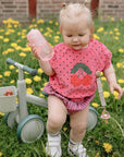 Toddler in pink cherry shirt on balance bike with FRIGG Daisy rubber pacifier, among dandelions.