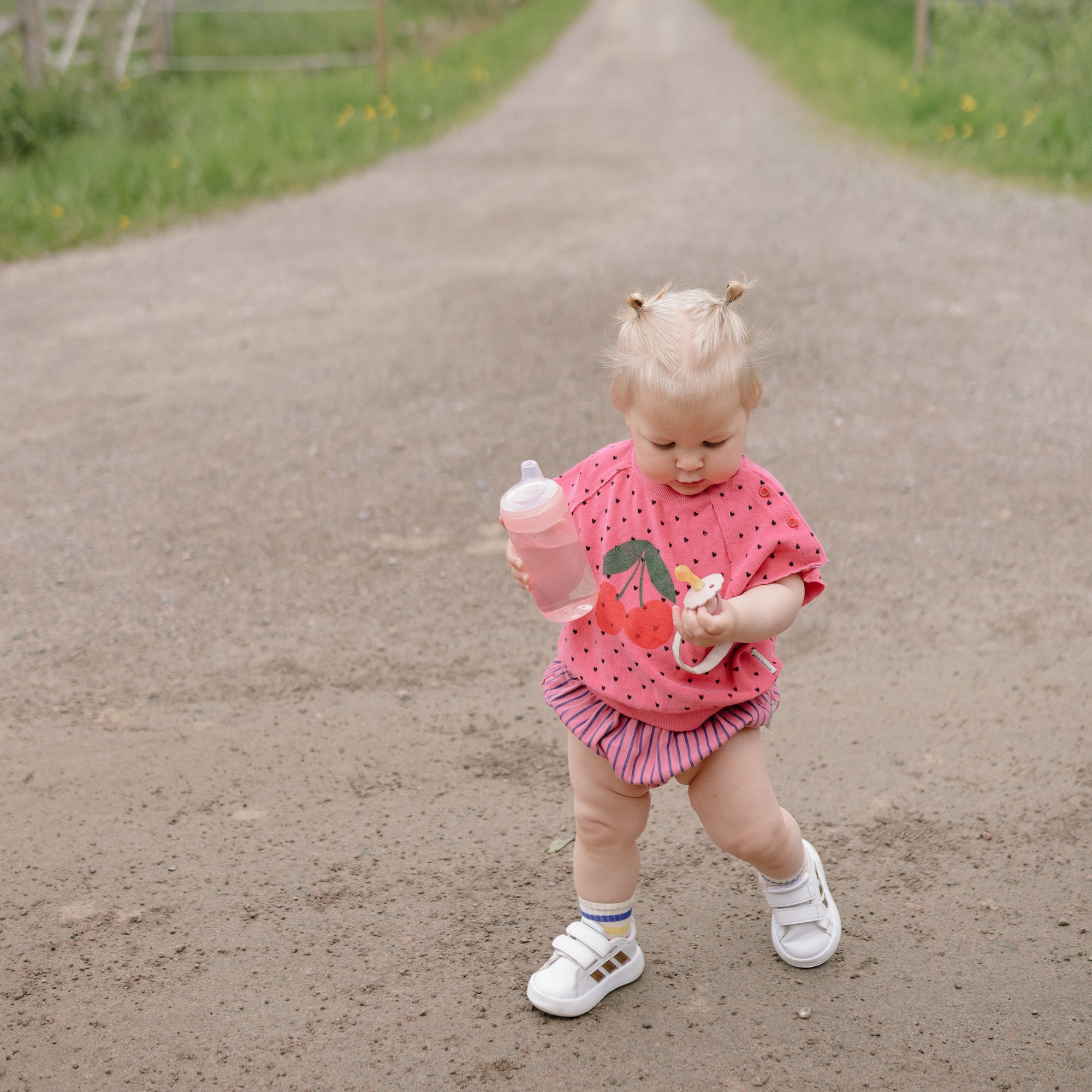 Toddler in pink wearing FRIGG Daisy Natural Rubber Pacifier, walking on a gravel path outside.