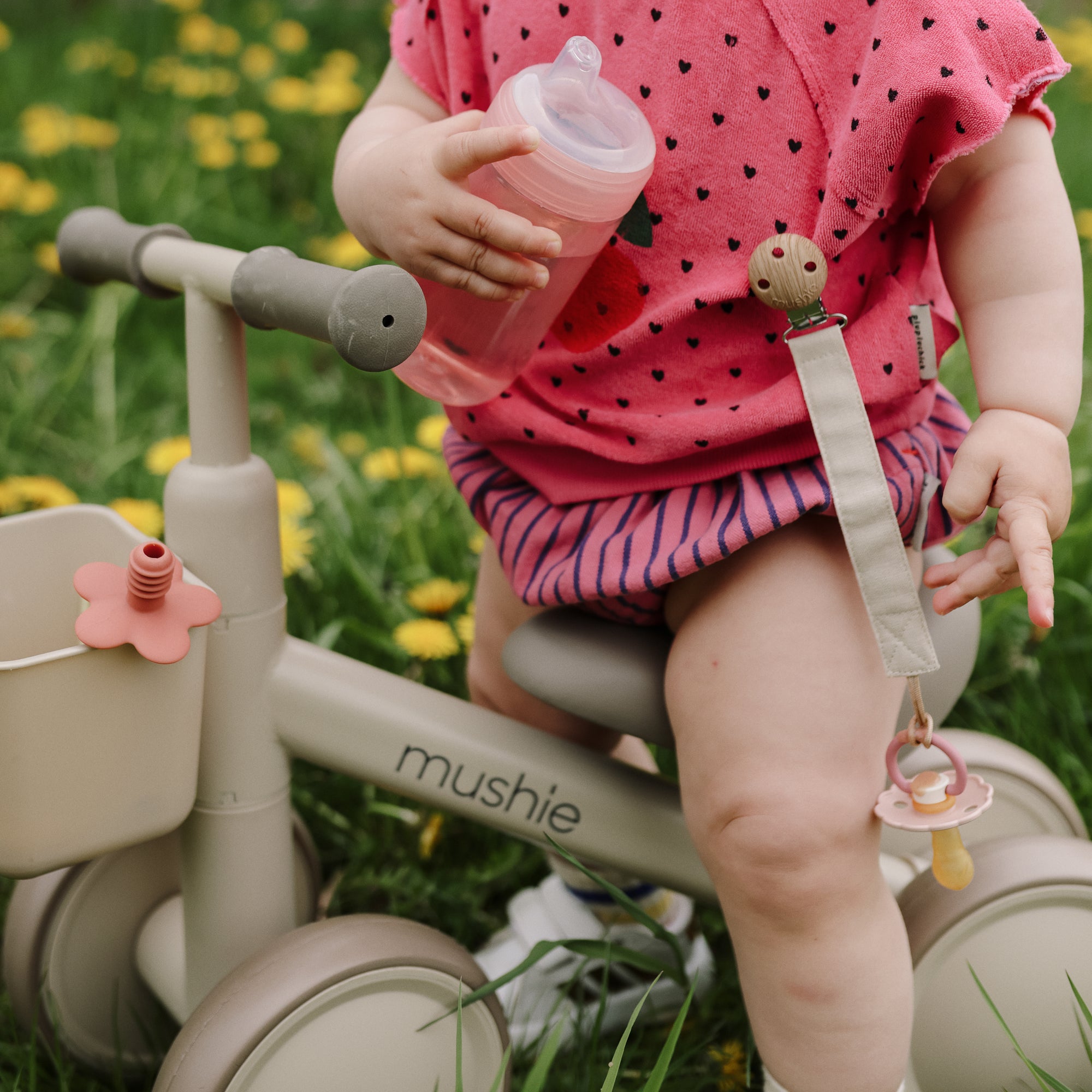 Toddler on a tricycle holding a mushie Soft Spout Sippy Cup, with a pacifier clipped on.