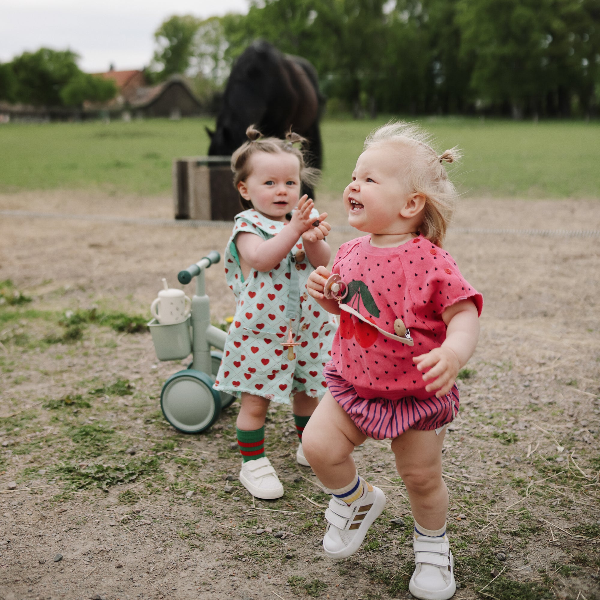 Two toddlers play outside, each holding a FRIGG Daisy Natural Rubber Pacifier 2-Pack.