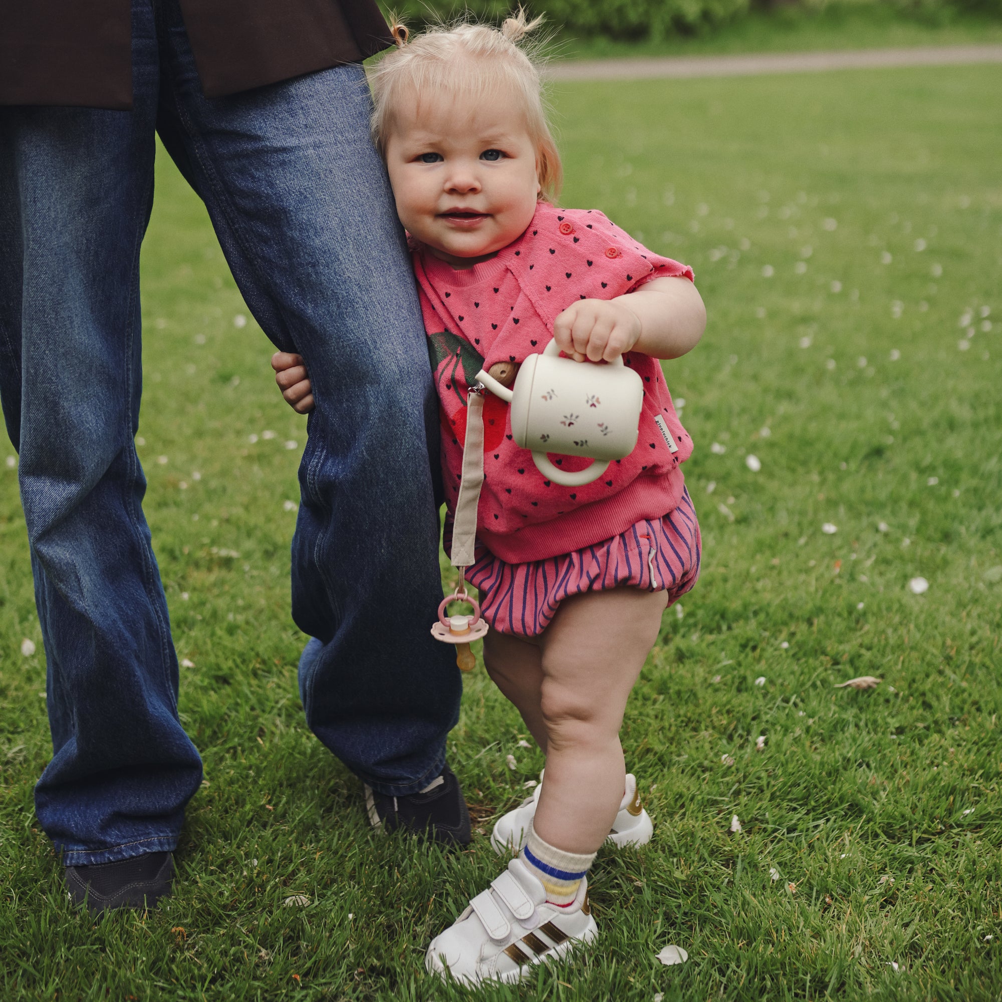 Toddler with FRIGG Daisy Natural Rubber Pacifier beside adult on green grass outdoors.
