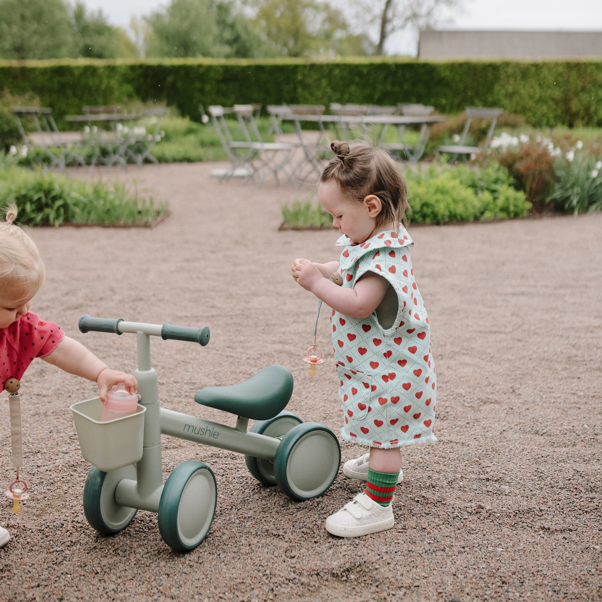 Two toddlers ride a green mushie Mini Toddler Bike on a gravel path in the garden.