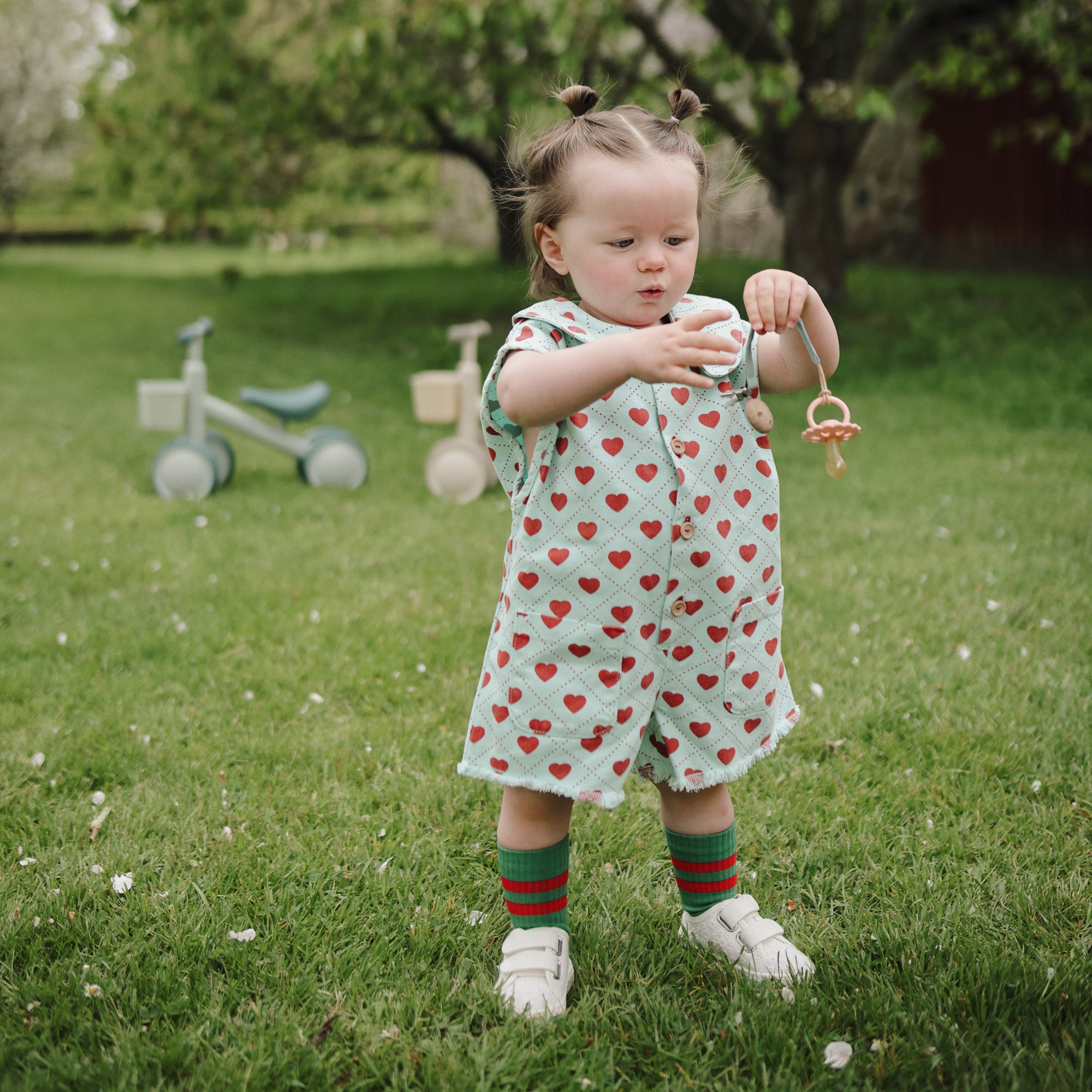 Toddler with mushie Linen Pacifier Clip 2-Pack holds pacifier while playing outdoors.