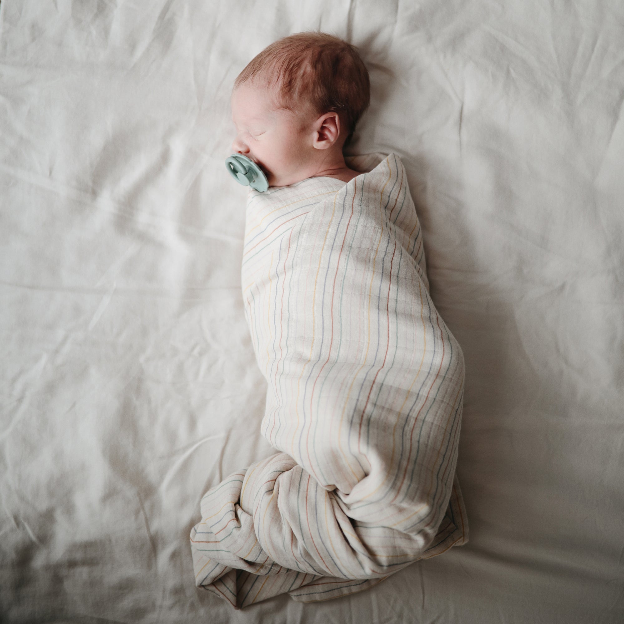 Newborn wrapped in mushie Organic Cotton Muslin Swaddle Blanket, lying with pacifier.