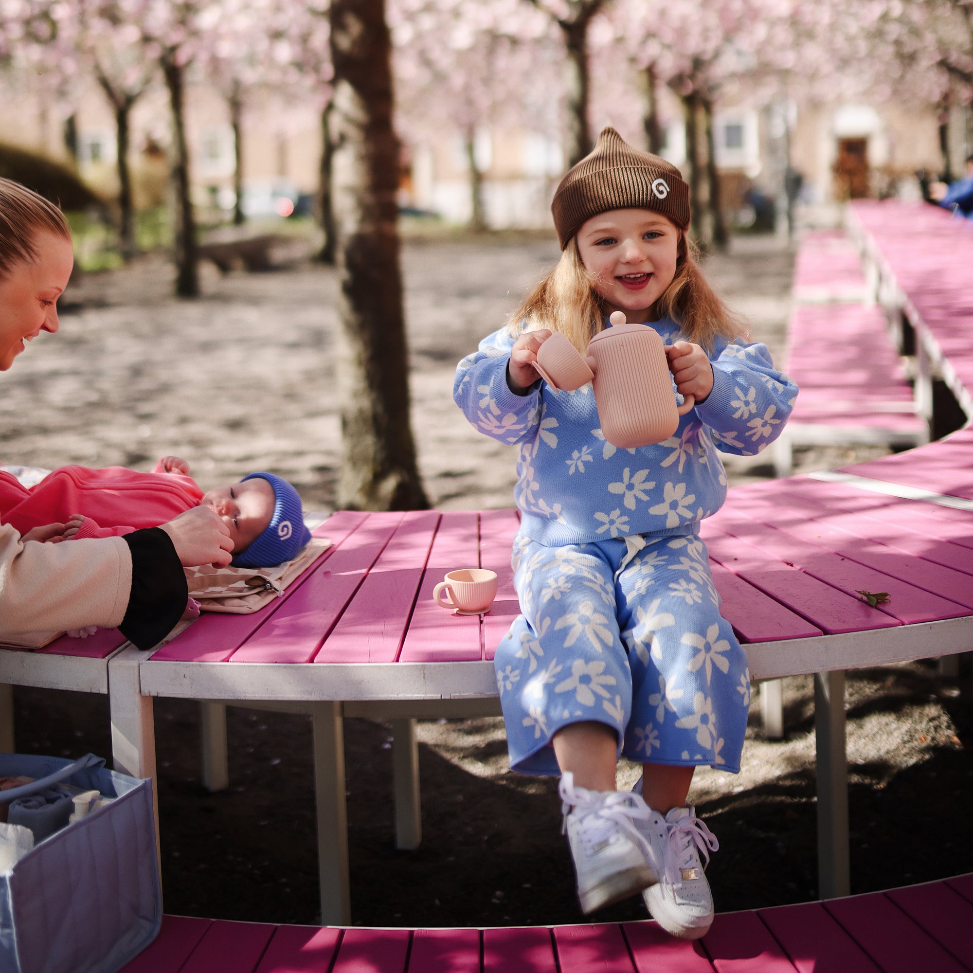 Young girl pours tea from Mushie's Silicone Tea Play Set at a pink table under blooming trees.
