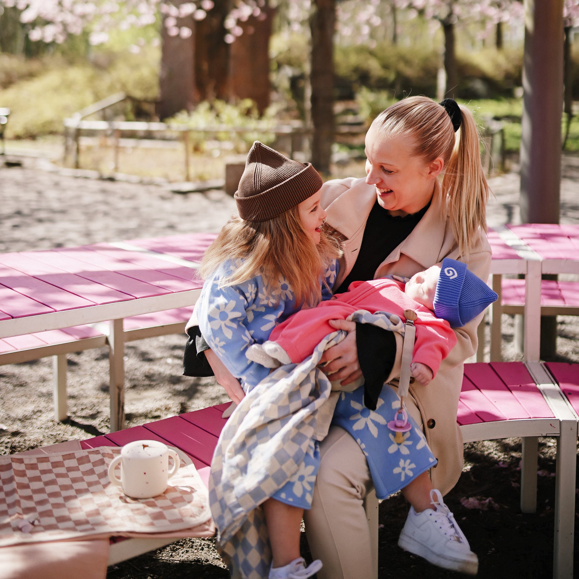 A woman holds two kids with mushie Linen Pacifier Clip 2-Pack at a pink picnic table.