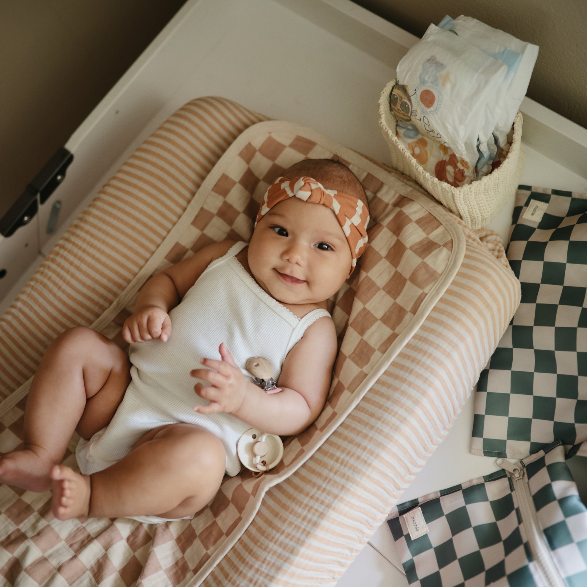 Smiling baby with mushie Linen Pacifier Clip 2-Pack on a checkered changing pad, diapers nearby.