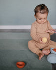 Baby in beige on rug plays with yellow ball and mushie Nesting Spheres Sensory Toy.