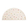 The mushie Silicone Placemat is cream-colored with a half-circle shape, made from food-grade silicone, and features small polka dots in red, yellow, green, and blue. "mushie" is embossed in the bottom right corner.