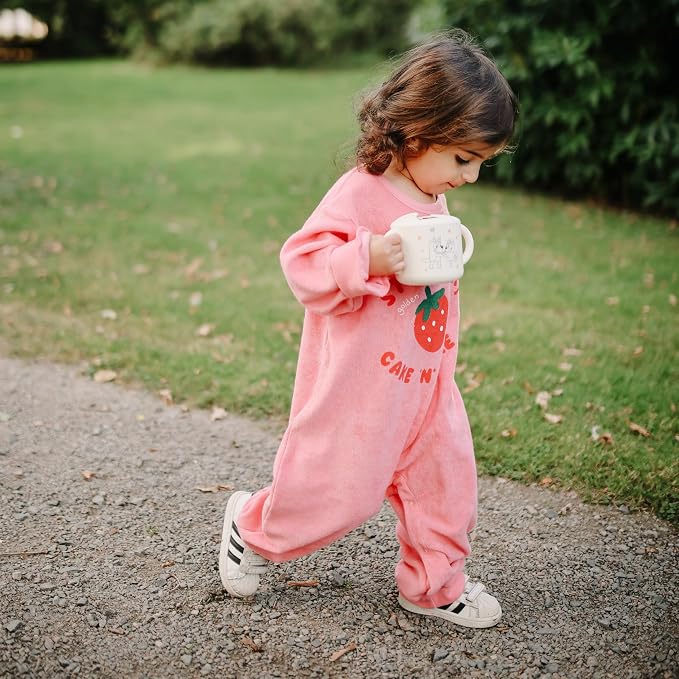 A toddler girl walking outdoors on a gravel path wearing a pink long-sleeve one-piece jumpsuit with a strawberry graphic and the text 'CAKE N' on the front, holding a white mug.