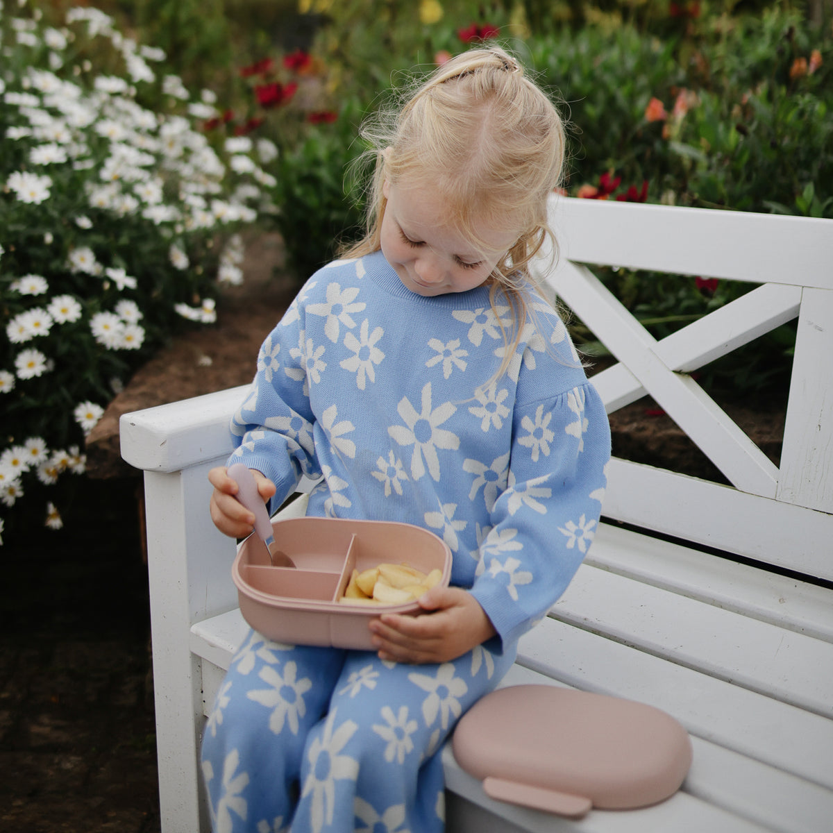 A child sits on a bench holding an open mushie Lunchbox with a spoon.