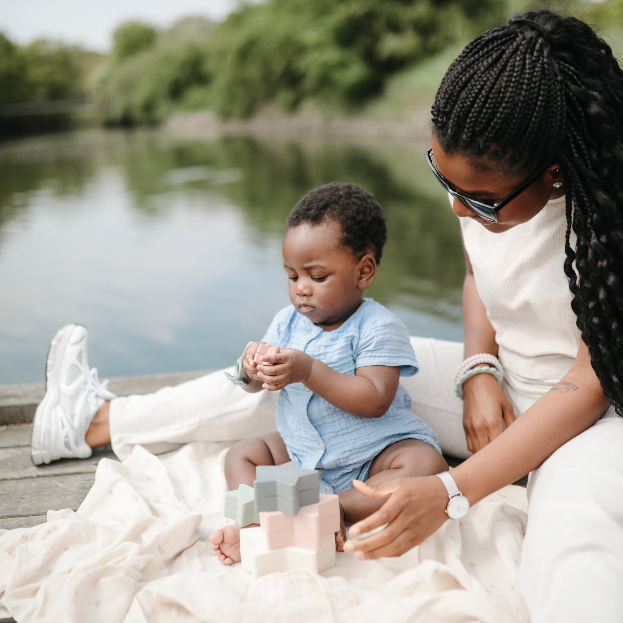 A woman and baby play with mushie Nesting Stars Toy on a blanket by a lake.