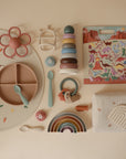 A flat lay of baby items in soft pastels, featuring a divided silicone plate, silicone utensils, stacking toys, teether, pacifier, rainbow toy, muslin cloth, soft textured toy, and the mushie Reusable Sticker Set.