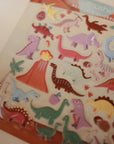 The mushie Reusable Sticker Set features colorful cartoon dinosaurs, a volcano, palm trees, and dino eggs on a beige background—perfect for fans of reusable sticker sets or sticker books.