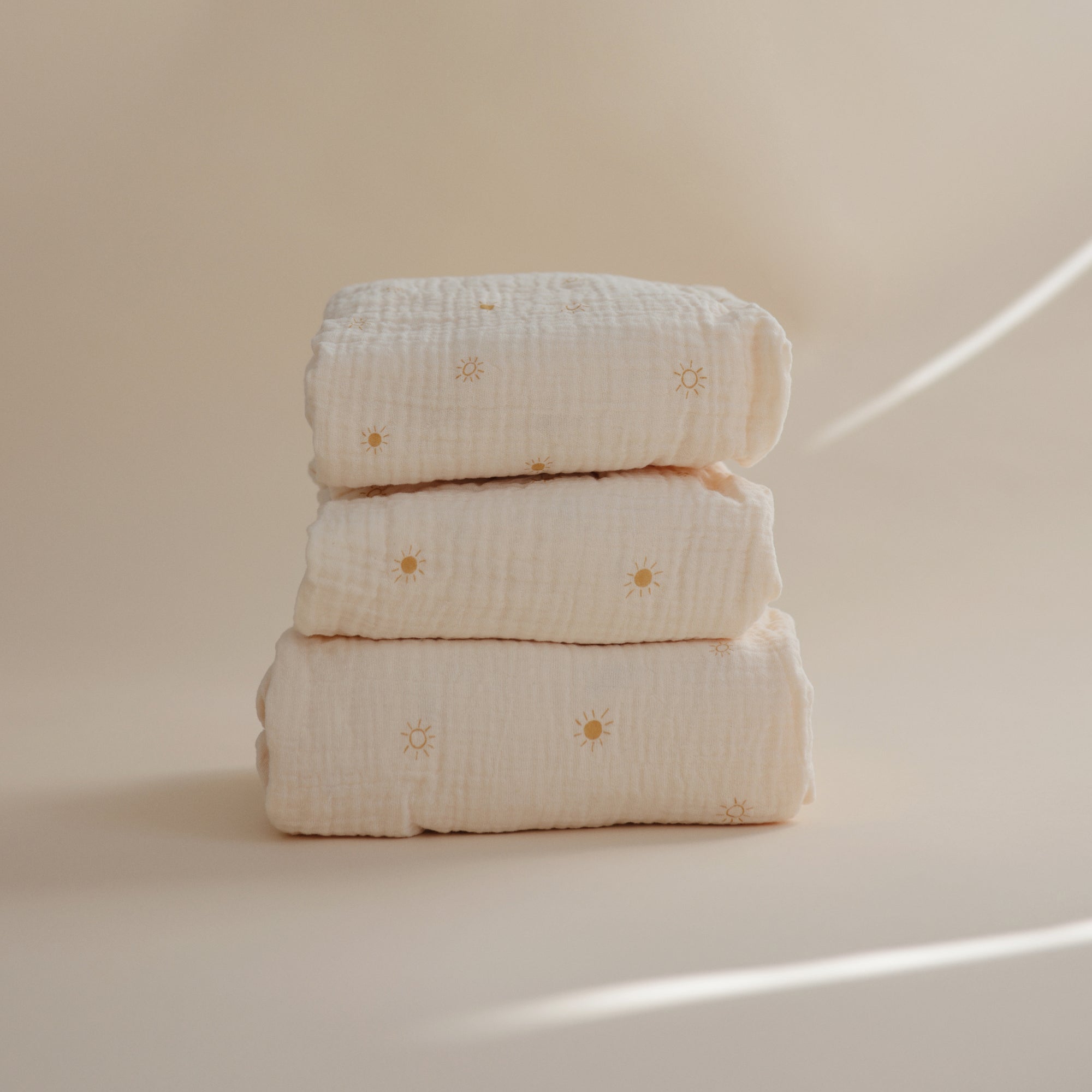 Three mushie Extra Soft Muslin Crib Sheets, cream-colored, stacked on a neutral sun-patterned background.