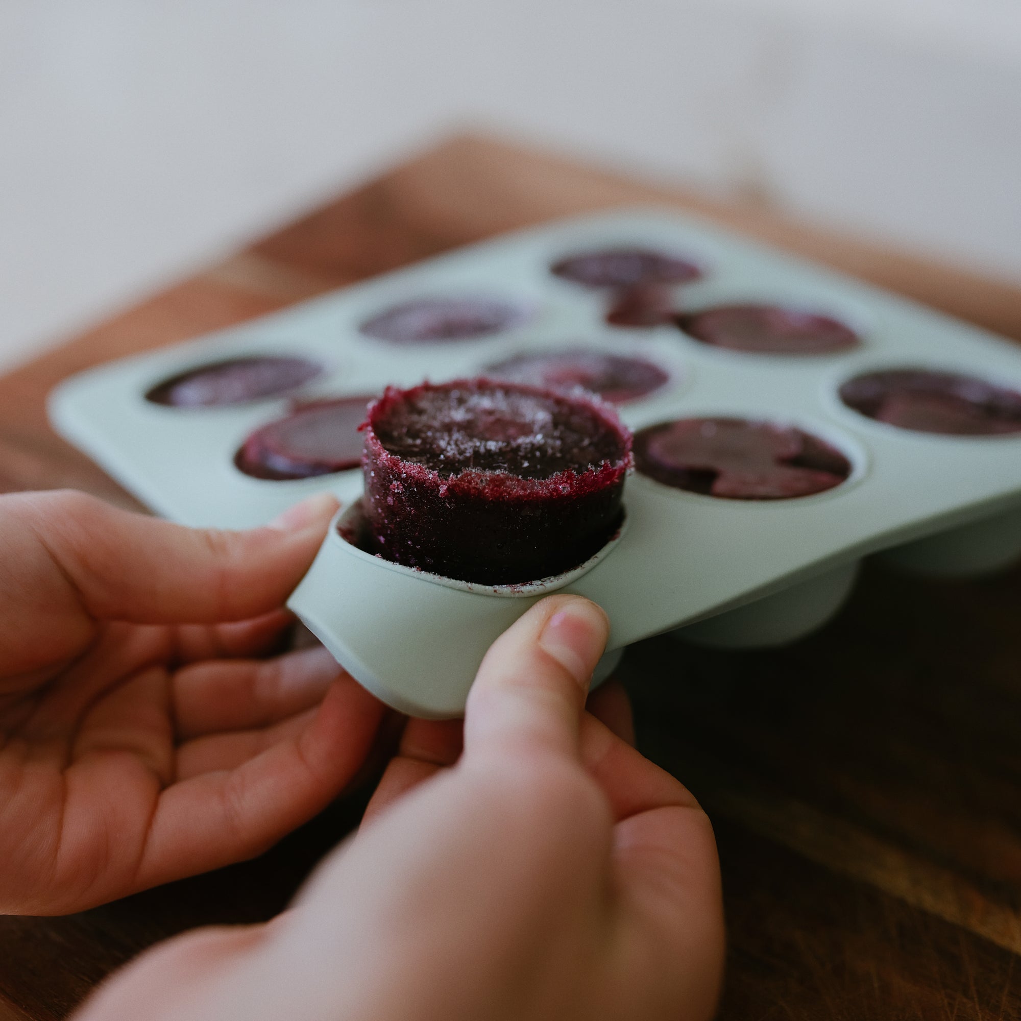 Hands removing a frozen berry mix from a Mushie Baby Food Freezer Tray on a wooden surface.