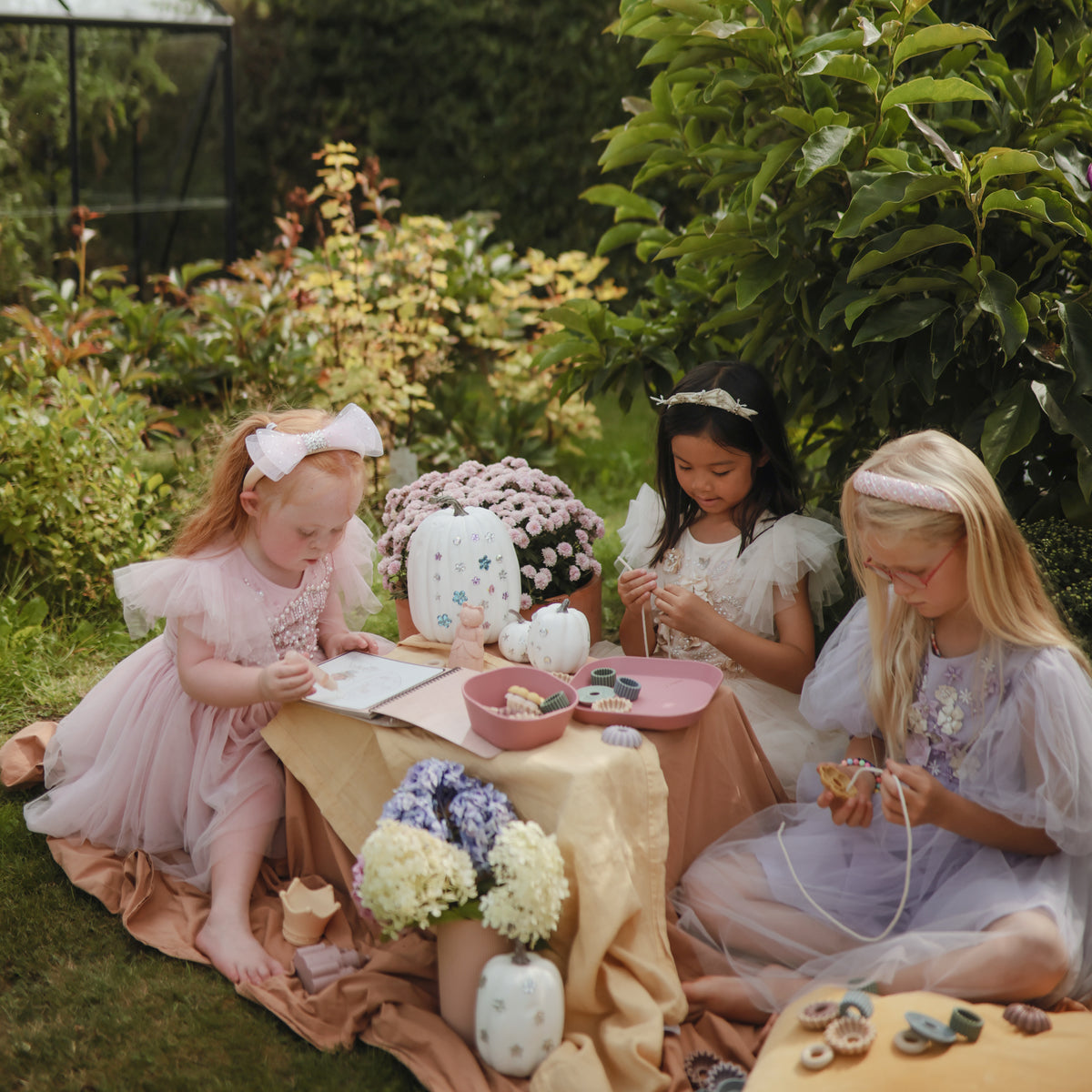 Three girls have a tea party outdoors, painting with mushie’s Reusable Water Color Pad amid flowers.