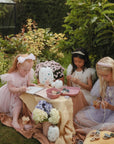Three girls have a tea party outdoors, painting with mushie’s Reusable Water Color Pad amid flowers.
